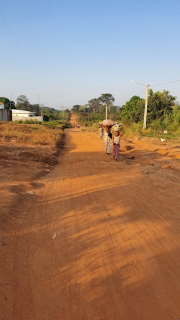 A rural dirt road stretches into the distance under a clear blue sky. Several people, including women in traditional clothing carrying items on their heads, walk along the road. The area is lush with green trees and vegetation lining the edges of the road, while utility poles follow the path. A white building is visible on the left, contrasting with the natural surroundings.