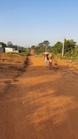 A rural dirt road stretches into the distance under a clear blue sky. Several people, including women in traditional clothing carrying items on their heads, walk along the road. The area is lush with green trees and vegetation lining the edges of the road, while utility poles follow the path. A white building is visible on the left, contrasting with the natural surroundings.