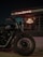 A motorcycle is parked in front of a building with illuminated signage. The building has a large visible window displaying the inner workings of a workshop or showroom. Prominent branding suggests it is associated with a particular motorcycle brand.