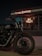 A motorcycle is parked in front of a building with illuminated signage. The building has a large visible window displaying the inner workings of a workshop or showroom. Prominent branding suggests it is associated with a particular motorcycle brand.