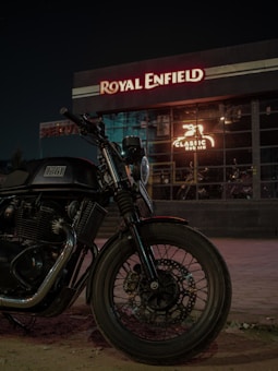 A motorcycle is parked in front of a building with illuminated signage. The building has a large visible window displaying the inner workings of a workshop or showroom. Prominent branding suggests it is associated with a particular motorcycle brand.