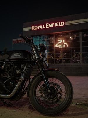 A motorcycle is parked in front of a building with illuminated signage. The building has a large visible window displaying the inner workings of a workshop or showroom. Prominent branding suggests it is associated with a particular motorcycle brand.
