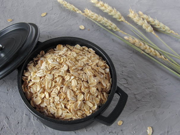 A black bowl filled with rolled oats is placed on a textured gray surface. Beside the bowl, there are several stalks of wheat laid out.