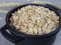 A black bowl filled with uncooked rolled oats, placed on a textured gray surface. In the background, there are wheat stalks slightly out of focus.