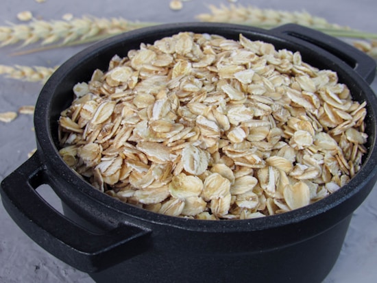 A black bowl filled with uncooked rolled oats, placed on a textured gray surface. In the background, there are wheat stalks slightly out of focus.