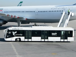 An airport scene featuring a large commercial airplane on the tarmac. A passenger stairs vehicle is attached to the plane, with a person standing near the top of the stairs. In the foreground, an airport bus labeled 'Cobus 3000' and 'IranAir' is parked parallel to the airplane.