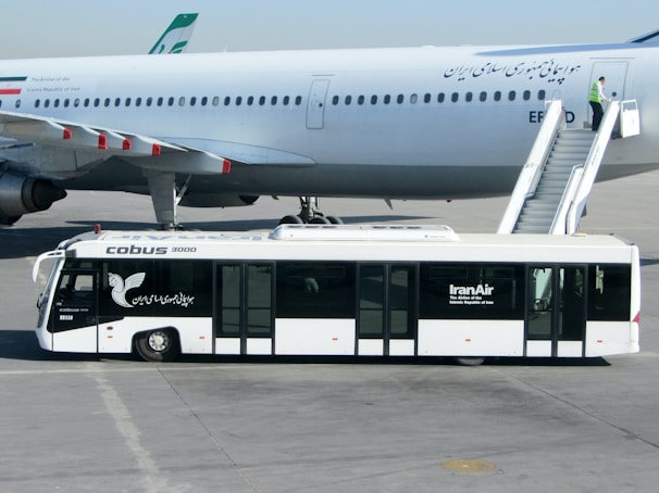 An airport scene featuring a large commercial airplane on the tarmac. A passenger stairs vehicle is attached to the plane, with a person standing near the top of the stairs. In the foreground, an airport bus labeled 'Cobus 3000' and 'IranAir' is parked parallel to the airplane.