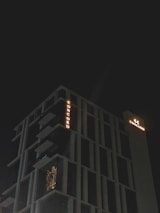 A modern high-rise building with illuminated signage displaying the word 'CONCORD' against a dark night sky. The structure features distinct geometric architectural elements and balconies. There is a warm glow emanating from one of the windows, suggesting interior lighting.