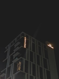 A modern high-rise building with illuminated signage displaying the word 'CONCORD' against a dark night sky. The structure features distinct geometric architectural elements and balconies. There is a warm glow emanating from one of the windows, suggesting interior lighting.