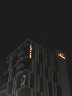 A modern high-rise building with illuminated signage displaying the word 'CONCORD' against a dark night sky. The structure features distinct geometric architectural elements and balconies. There is a warm glow emanating from one of the windows, suggesting interior lighting.
