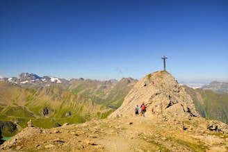 A group of hikers is making their way to the peak of a rocky mountain, where a large cross is visible. The scene is set against a backdrop of distant mountain ranges and clear blue skies. The landscape features rugged terrain and sparse vegetation.