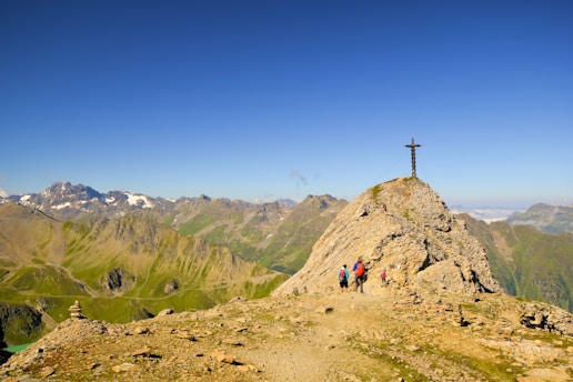 A group of hikers is making their way to the peak of a rocky mountain, where a large cross is visible. The scene is set against a backdrop of distant mountain ranges and clear blue skies. The landscape features rugged terrain and sparse vegetation.