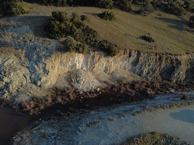A rocky hillside with visible erosion patterns, surrounded by grassy terrain and patches of dense, dark green trees. A small stream or creek flows at the bottom, bordered by rocky and sandy areas.