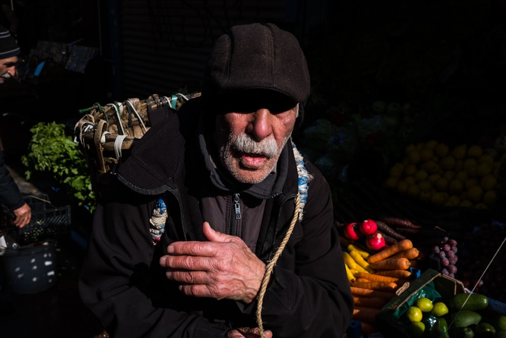 a man in a black jacket and hat standing in front of a bunch of vegetables