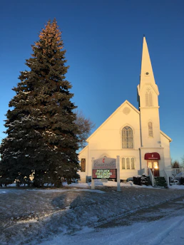 A quaint white church with a tall steeple is set against a clear blue sky. A large pine tree stands to the left of the church. There is snow on the ground, adding to the calm winter atmosphere. The church sign reads 'Casenville United Methodist Church' with an announcement for a potluck following worship.