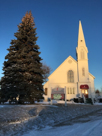 A quaint white church with a tall steeple is set against a clear blue sky. A large pine tree stands to the left of the church. There is snow on the ground, adding to the calm winter atmosphere. The church sign reads 'Casenville United Methodist Church' with an announcement for a potluck following worship.