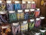 An assortment of glass jars filled with colorful candy sticks, each labeled with different flavors such as blackberry, blueberry, lemon, and licorice. The jars are neatly arranged on wooden shelves in a display that highlights their vibrant hues.