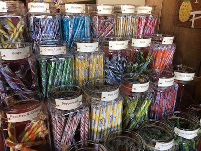 An assortment of glass jars filled with colorful candy sticks, each labeled with different flavors such as blackberry, blueberry, lemon, and licorice. The jars are neatly arranged on wooden shelves in a display that highlights their vibrant hues.