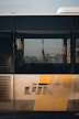 A close-up view of the side of a De Lijn bus with its logo prominently displayed. The bus window reflects some outdoor scenes, likely an urban environment.