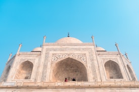 A majestic white marble building with intricate carvings and large arched doorways rises against a clear blue sky. The architecture features detailed floral patterns and decorative elements. In the foreground, people can be seen standing near the central arch.