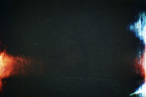 A dark background punctuated by abstract splashes of red and blue light on opposite sides. The left side features a warm red glow, while the right side displays cooler blue tones, merging into hints of white.