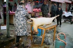 A street market setting features a man in a camouflaged jacket tending to a large animal carcass lying on a wooden table. Other individuals, mostly older adults, are observing the scene. Various market stalls in the background display signs and meat products. An empty red and blue gas cylinder is situated nearby on the pavement, and the atmosphere suggests a bustling, open-air marketplace.