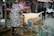 A street market setting features a man in a camouflaged jacket tending to a large animal carcass lying on a wooden table. Other individuals, mostly older adults, are observing the scene. Various market stalls in the background display signs and meat products. An empty red and blue gas cylinder is situated nearby on the pavement, and the atmosphere suggests a bustling, open-air marketplace.