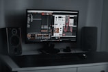 A desktop computer setup featuring a monitor displaying a digital audio workstation with modular synthesizer software. Dual speakers are positioned on either side of the monitor. A keyboard and mouse are placed on a sleek, dark desk.