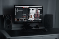 A desktop computer setup featuring a monitor displaying a digital audio workstation with modular synthesizer software. Dual speakers are positioned on either side of the monitor. A keyboard and mouse are placed on a sleek, dark desk.