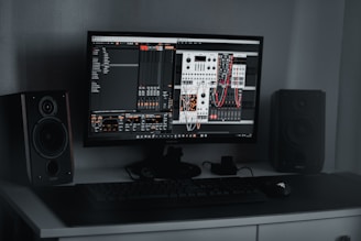 A desktop computer setup featuring a monitor displaying a digital audio workstation with modular synthesizer software. Dual speakers are positioned on either side of the monitor. A keyboard and mouse are placed on a sleek, dark desk.