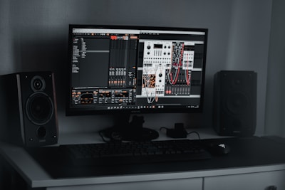 A desktop computer setup featuring a monitor displaying a digital audio workstation with modular synthesizer software. Dual speakers are positioned on either side of the monitor. A keyboard and mouse are placed on a sleek, dark desk.