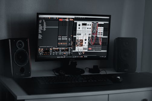 A desktop computer setup featuring a monitor displaying a digital audio workstation with modular synthesizer software. Dual speakers are positioned on either side of the monitor. A keyboard and mouse are placed on a sleek, dark desk.