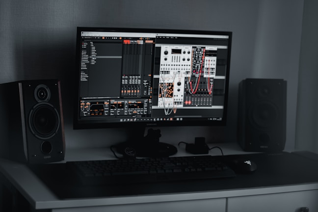 A desktop computer setup featuring a monitor displaying a digital audio workstation with modular synthesizer software. Dual speakers are positioned on either side of the monitor. A keyboard and mouse are placed on a sleek, dark desk.