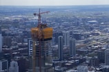 A cityscape featuring a skyscraper under construction with a crane atop, surrounded by numerous buildings and a vast urban sprawl. The area appears densely populated with an array of architectural styles.