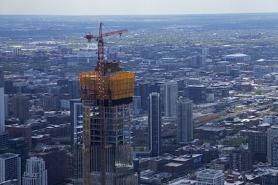 A cityscape featuring a skyscraper under construction with a crane atop, surrounded by numerous buildings and a vast urban sprawl. The area appears densely populated with an array of architectural styles.