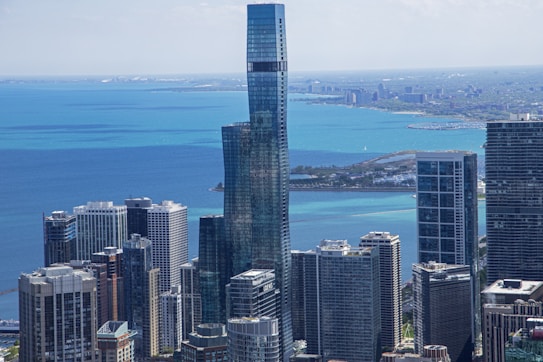A skyline view featuring modern skyscrapers with a prominent, uniquely curved glass building in the center. The background highlights a vast body of water with a gradient of blue hues and traces of urban development along the shoreline.