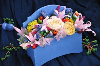 A vibrant floral arrangement featuring a variety of flowers in a decorative light blue vase. The bouquet includes pale yellow roses, pink lilies, small red berries, and purple flowers, surrounded by green foliage. The setting is against a dark blue fabric background, which enhances the vivid colors of the flowers.