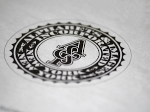 A circular black and white emblem or seal with the letter 'A' in the center. The surrounding text reads 'Alphabets & Images Incorporated'. The design features a detailed border and is set against a plain background.