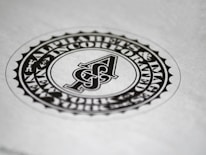 A circular black and white emblem or seal with the letter 'A' in the center. The surrounding text reads 'Alphabets & Images Incorporated'. The design features a detailed border and is set against a plain background.