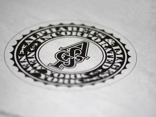 A circular black and white emblem or seal with the letter 'A' in the center. The surrounding text reads 'Alphabets & Images Incorporated'. The design features a detailed border and is set against a plain background.