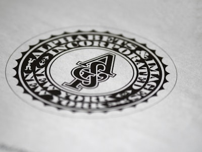A circular black and white emblem or seal with the letter 'A' in the center. The surrounding text reads 'Alphabets & Images Incorporated'. The design features a detailed border and is set against a plain background.