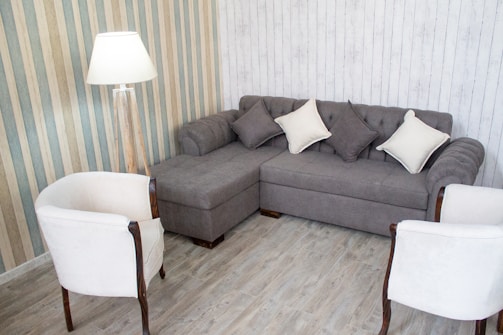 A cozy living room corner with a gray sectional sofa adorned with light and dark gray cushions. Two white armchairs with wooden accents face the sofa. A floor lamp with a white shade is positioned nearby. The walls display a mix of striped and wooden panel designs, and the flooring is a light wood laminate.