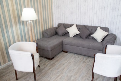 A cozy living room corner with a gray sectional sofa adorned with light and dark gray cushions. Two white armchairs with wooden accents face the sofa. A floor lamp with a white shade is positioned nearby. The walls display a mix of striped and wooden panel designs, and the flooring is a light wood laminate.