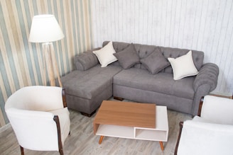 A cozy living room setup featuring a gray sectional sofa adorned with gray and white cushions. It is surrounded by two white armchairs with wooden arms. A wooden coffee table with a white shelf sits in the center. A standing lamp is positioned beside the sofa on a striped wallpapered wall, and the floor is covered with light-colored wood panels.