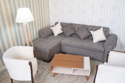 A cozy living room setup featuring a gray sectional sofa adorned with gray and white cushions. It is surrounded by two white armchairs with wooden arms. A wooden coffee table with a white shelf sits in the center. A standing lamp is positioned beside the sofa on a striped wallpapered wall, and the floor is covered with light-colored wood panels.