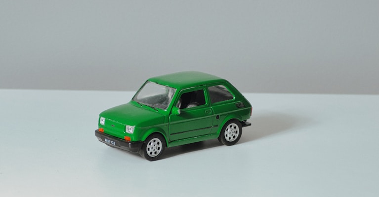 A small green toy car with a boxy design and black detailing sits on a flat, light-colored surface. The car features smooth lines, circular wheels with silver rims, and clear windows, giving it a retro look.