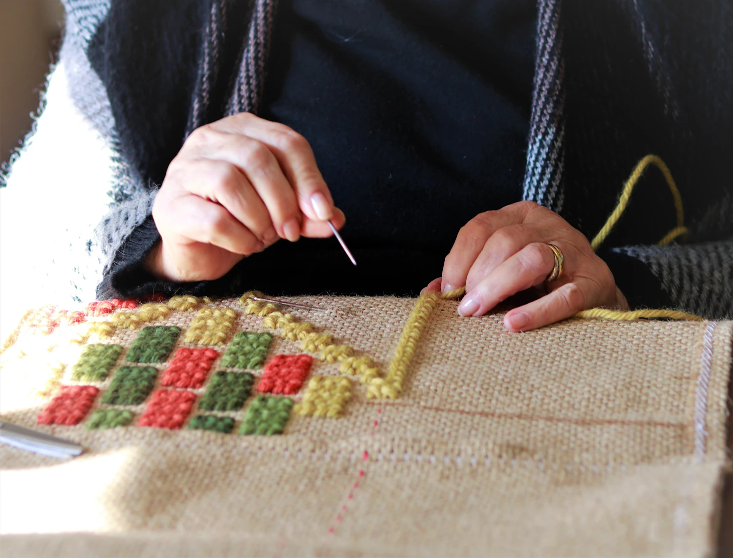 Two hands working a needle through heavy hessian webbing stretched across a bench
