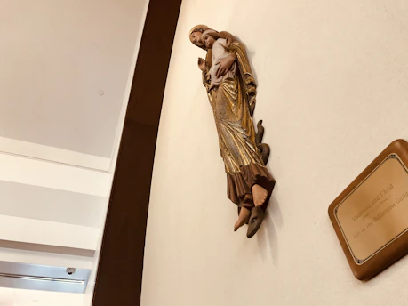 A statue of a woman holding a child is mounted on a wall. The woman is wearing a golden robe and is stepping on a snake. A plaque nearby reads 'Madonna and Child, Gift of the Rosary Shrine Guild'. The setting appears to be indoors with a white and brown color scheme.