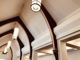 A series of elegant, arched ceiling beams made of dark wood with a polished finish. Suspended from the ceiling are modern, cylindrical pendant lights with a metal frame and frosted glass, emitting a soft, warm glow. The ceiling and walls are painted in neutral tones, enhancing the architectural details and providing a sense of spaciousness.