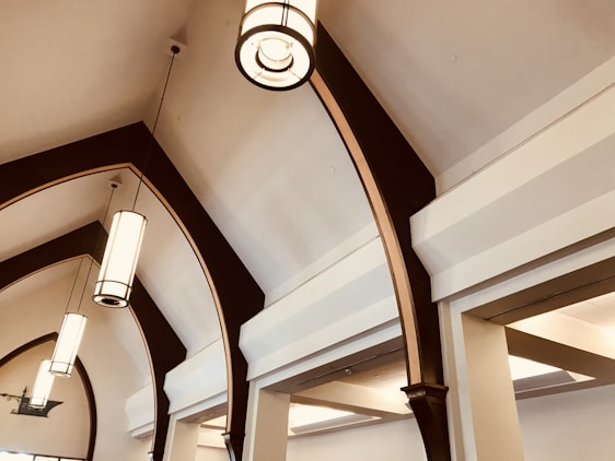 A series of elegant, arched ceiling beams made of dark wood with a polished finish. Suspended from the ceiling are modern, cylindrical pendant lights with a metal frame and frosted glass, emitting a soft, warm glow. The ceiling and walls are painted in neutral tones, enhancing the architectural details and providing a sense of spaciousness.